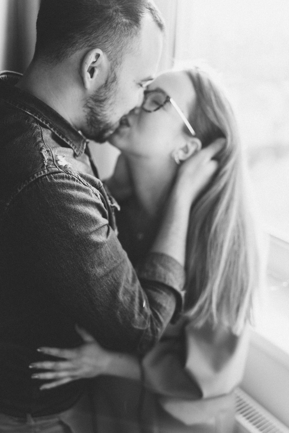 An intimate black and white photo of a romantic couple by a window.