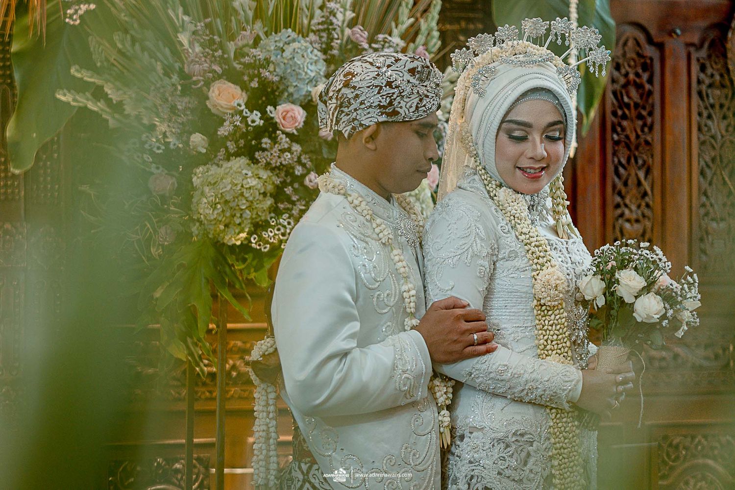 Elegant Indonesian wedding portrait with couple in traditional white attire against carved wooden backdrop.