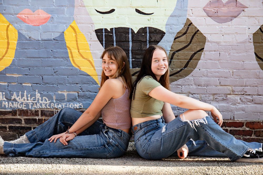 Two friends in casual outfits sitting back-to-back against colorful graffiti wall.
