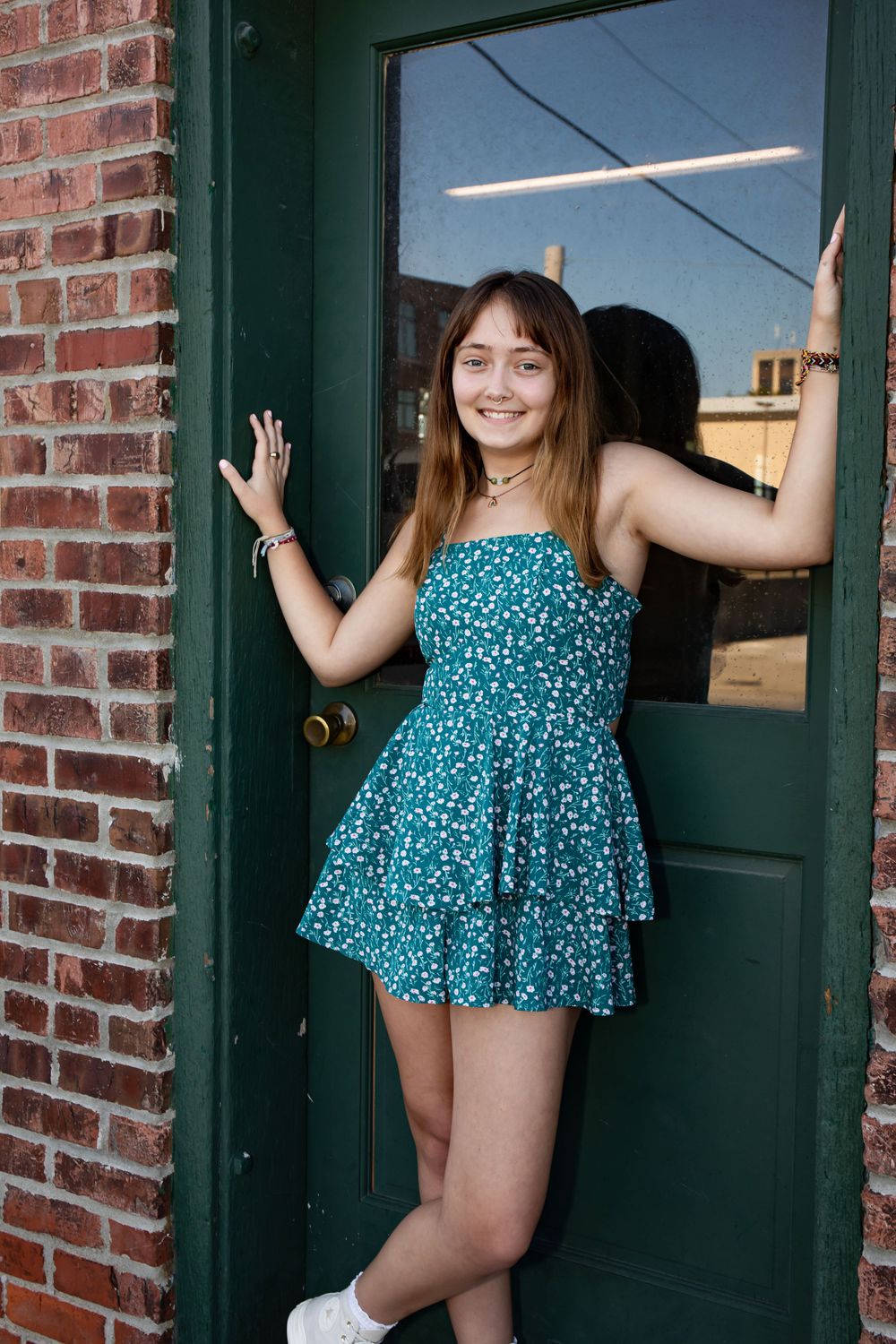 Person in a blue floral dress poses playfully by a green door against brick wall.
