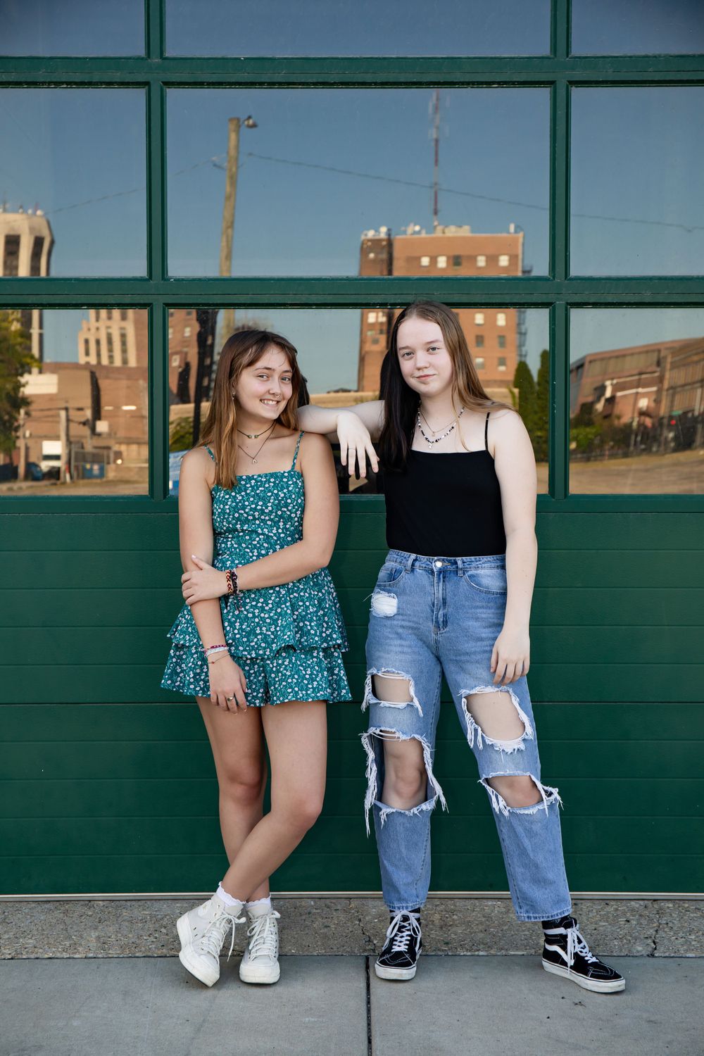 Two people pose against a green storefront window with urban buildings reflected in background.