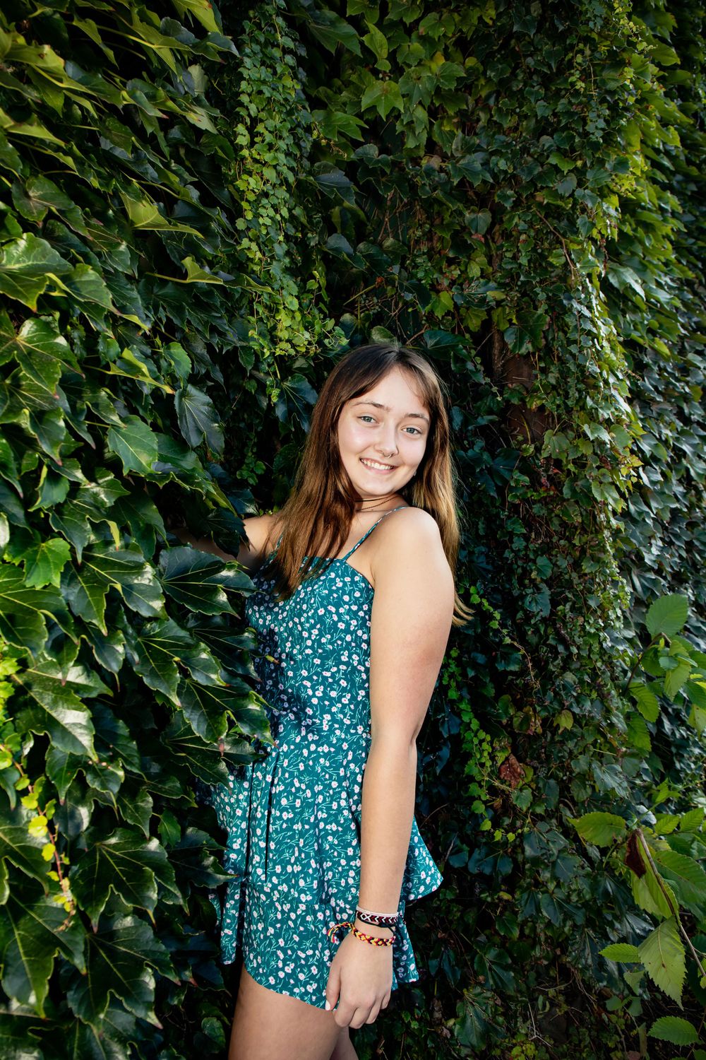 Person in teal dress poses against lush green ivy covered wall.