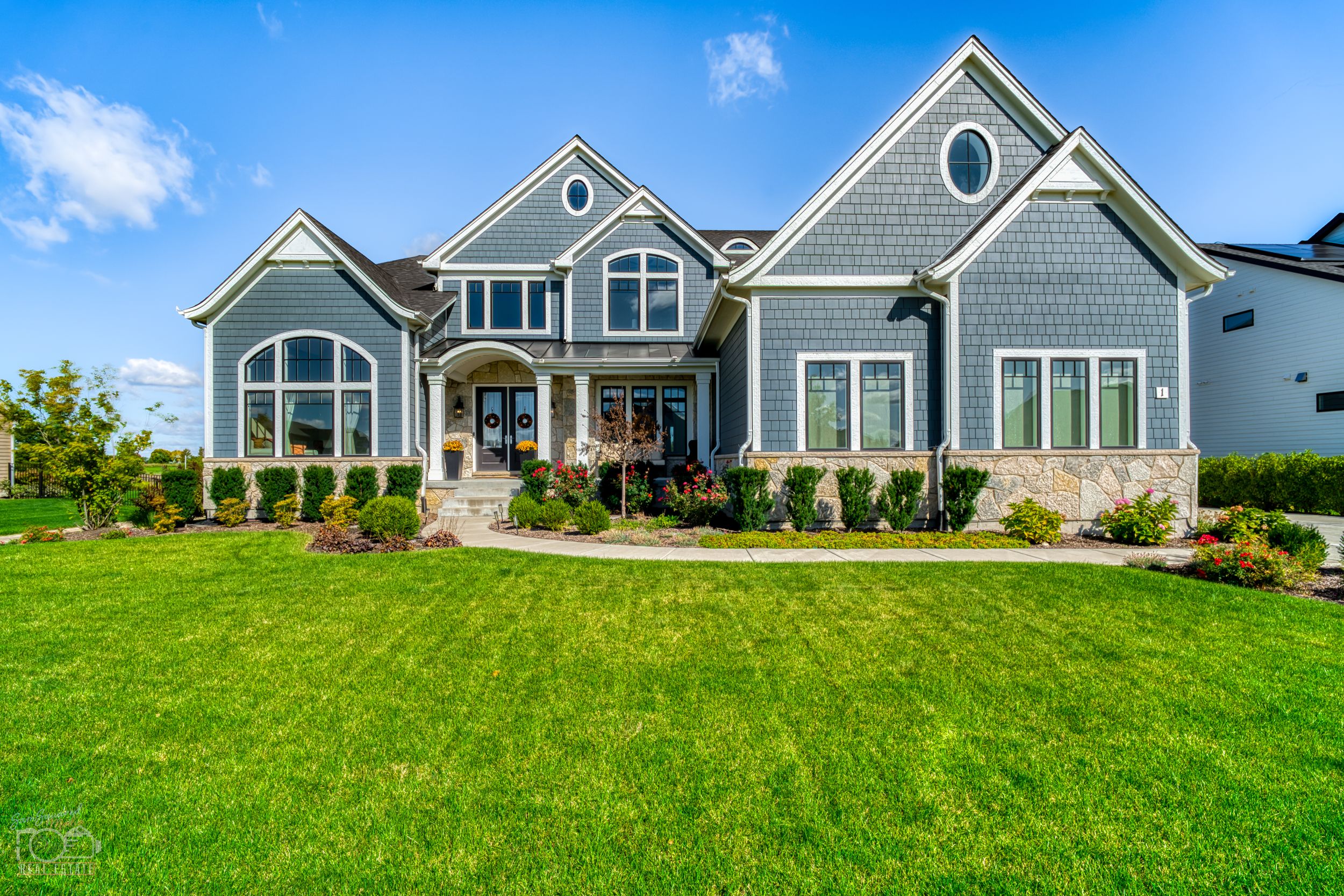 Gray craftsman style luxury home with beautiful landscaping and manicured lawn on sunny day.