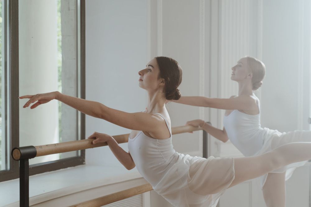 Dancers in white practicing arabesques at a ballet barre in a sunlit studio.