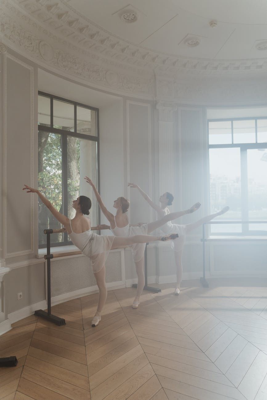 Ballet dancers practicing in formation at the barre in a bright classical studio space.