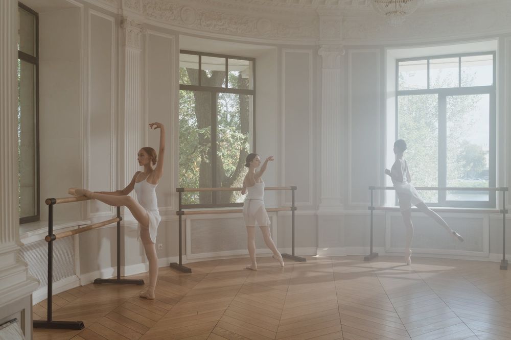 Ballet dancer practicing at barre in sunlit classical dance studio with wooden floors and ornate white walls.