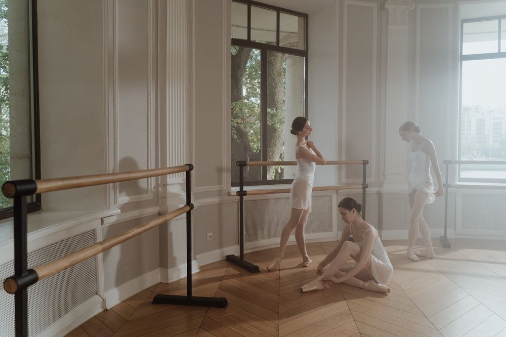 Dancers in white practicing various positions at the barre in a sunlit studio.
