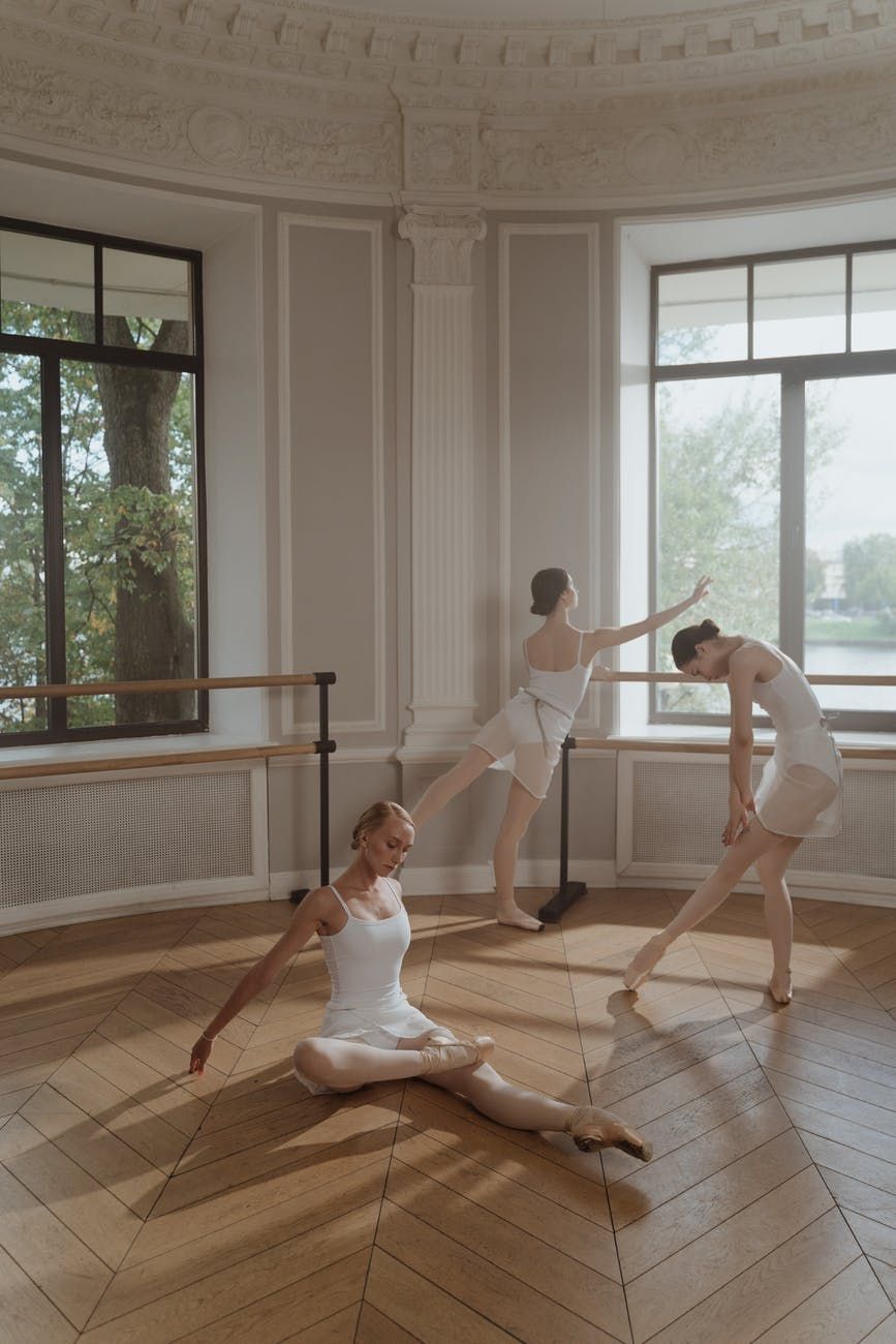 Ballet dancers in white practicing in an elegant studio with large windows and ornate moldings.