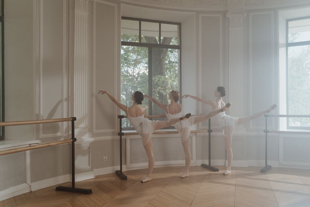 Dancers in white practicing arabesques at barre in bright classical studio with large windows.