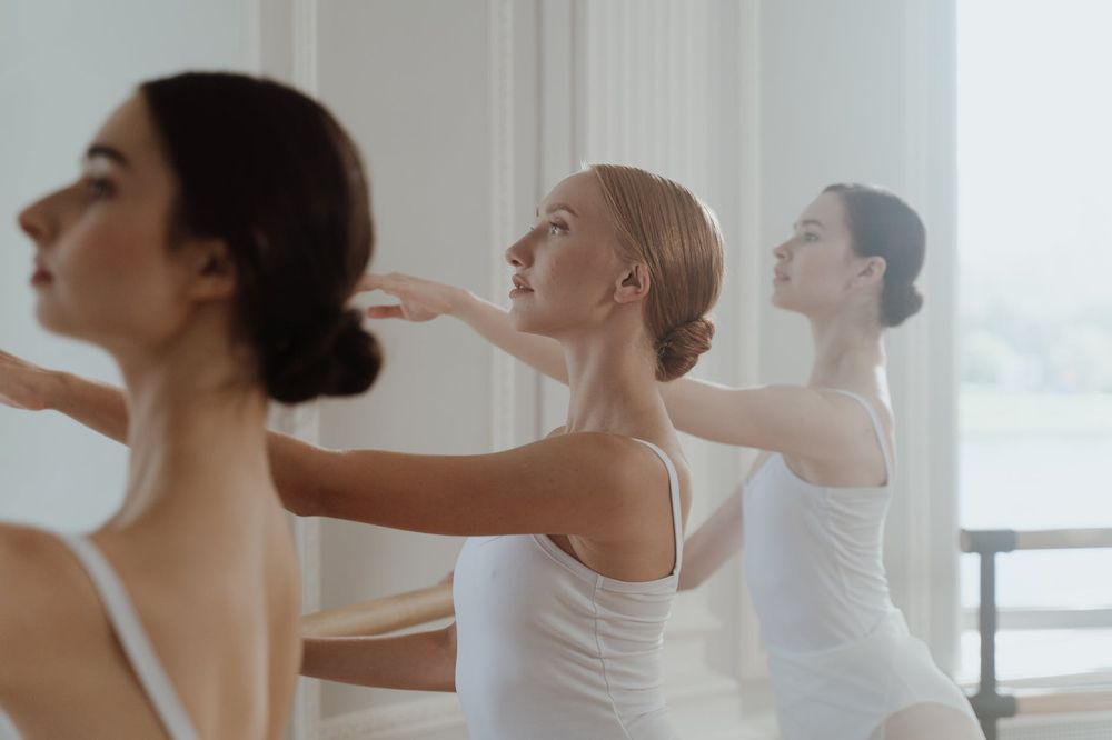 Dancers in white leotards practicing graceful arm movements during ballet class.
