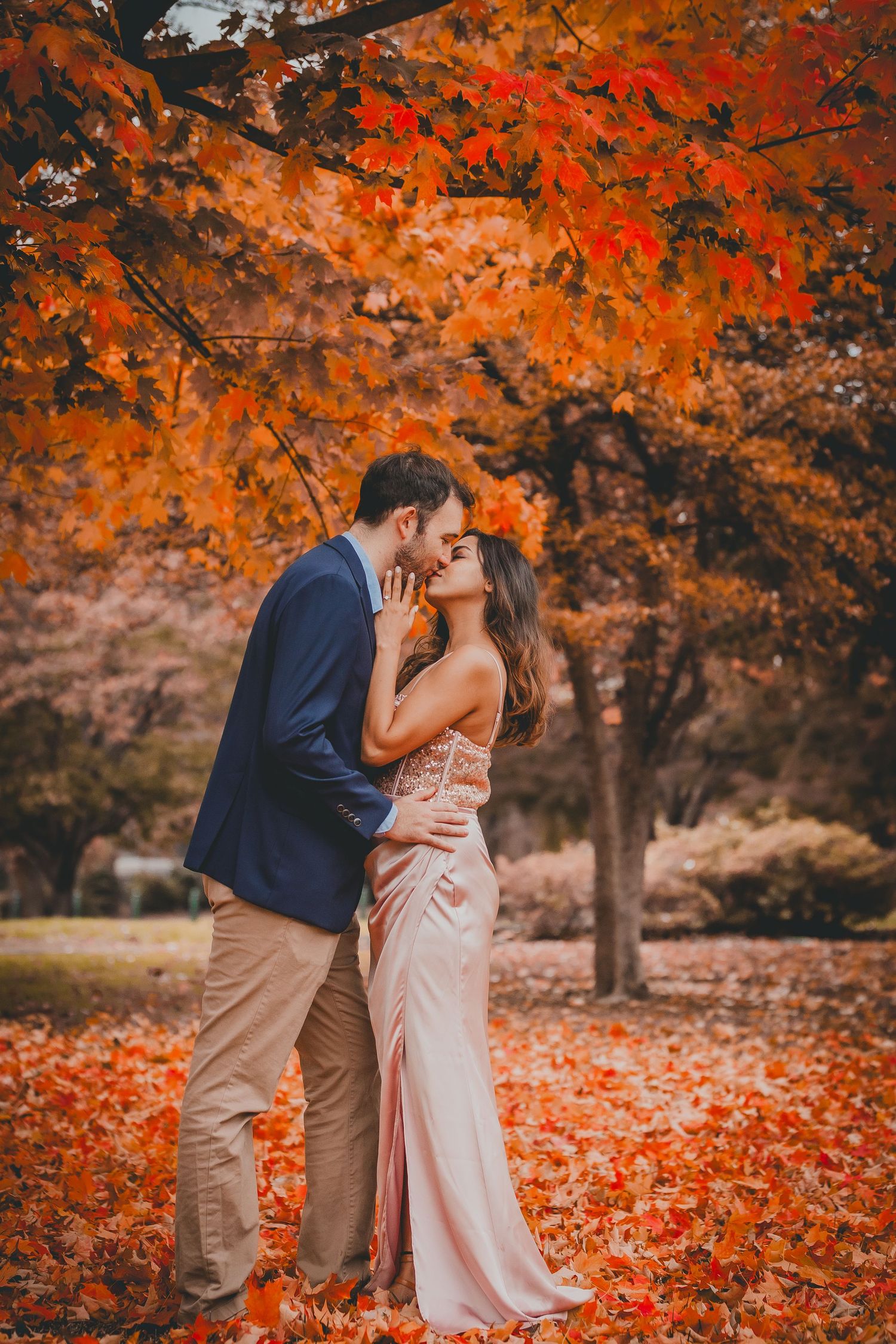 A couple embraces under vibrant orange autumn foliage with fallen leaves on the ground.