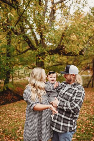 A couple in casual attire holds their small child while standing among fall trees.