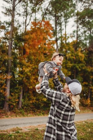 Person in plaid shirt lifts small child high in the air against backdrop of fall trees.