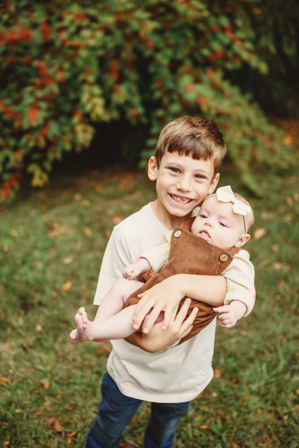 A young boy holds and smiles while holding a baby in a brown outfit against an autumn backdrop.