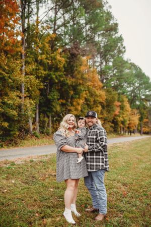 A couple embraces while standing on grass with colorful autumn trees in the background.