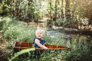 A happy baby sits in a wooden canoe surrounded by lush greenery near a stream.