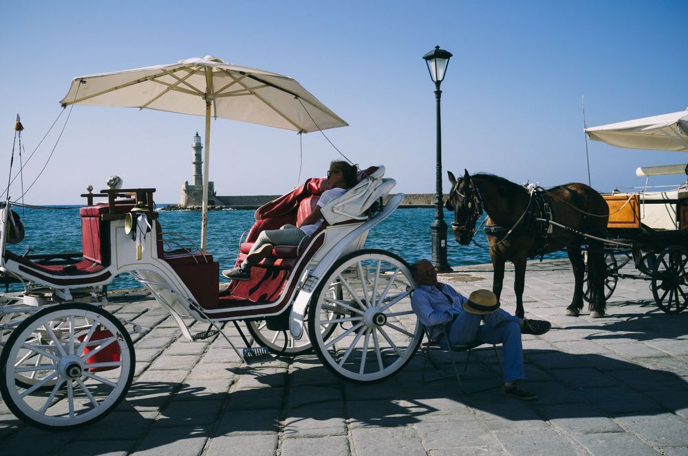 Horse-drawn carriages lined up along a sunny Mediterranean waterfront with beach umbrellas.
