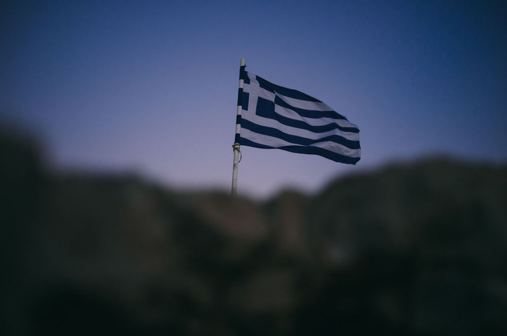 Greek flag waving against a dark mountainous landscape at dusk.