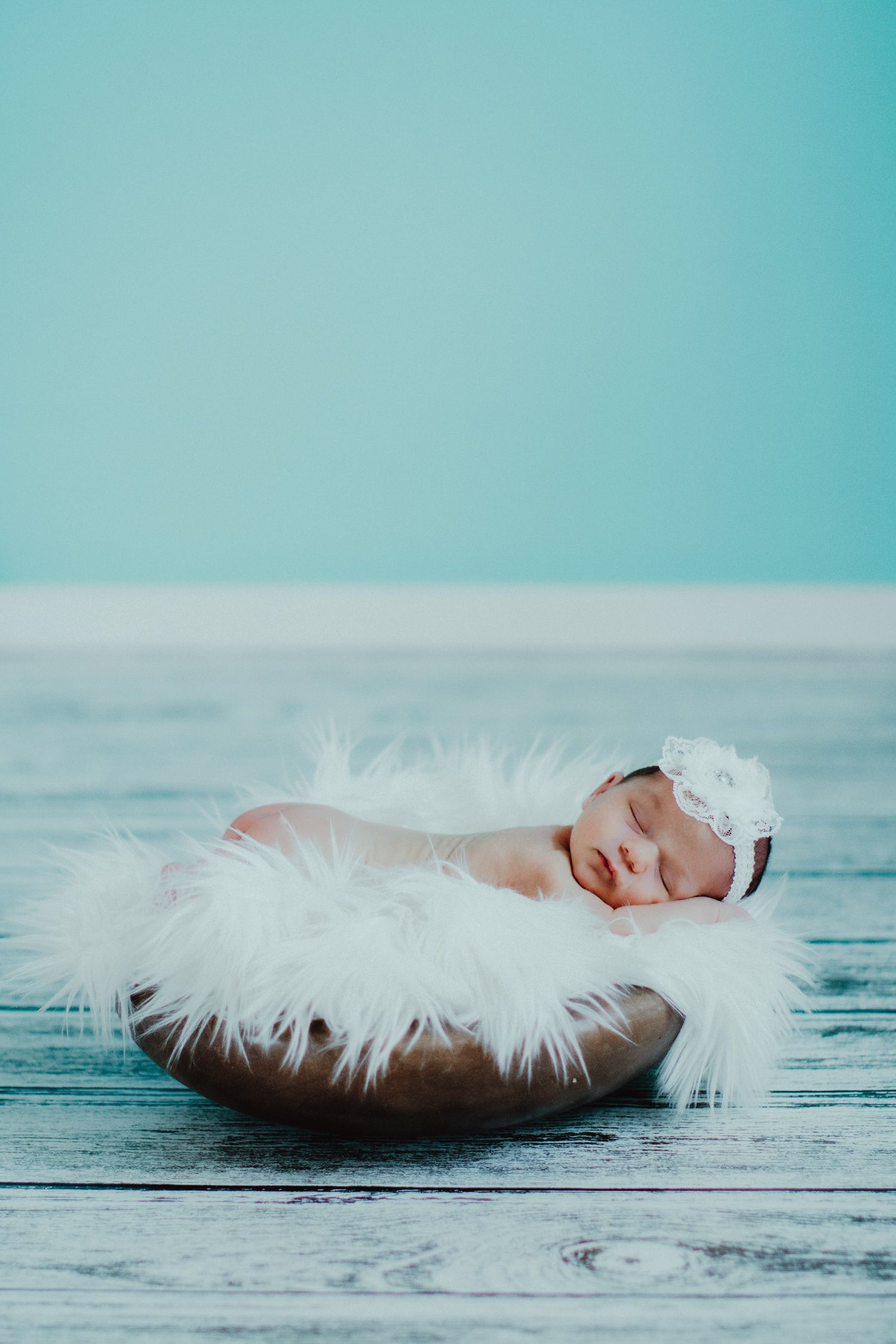 Newborn baby sleeping in a white fluffy nest on turquoise wooden backdrop with delicate white headband.