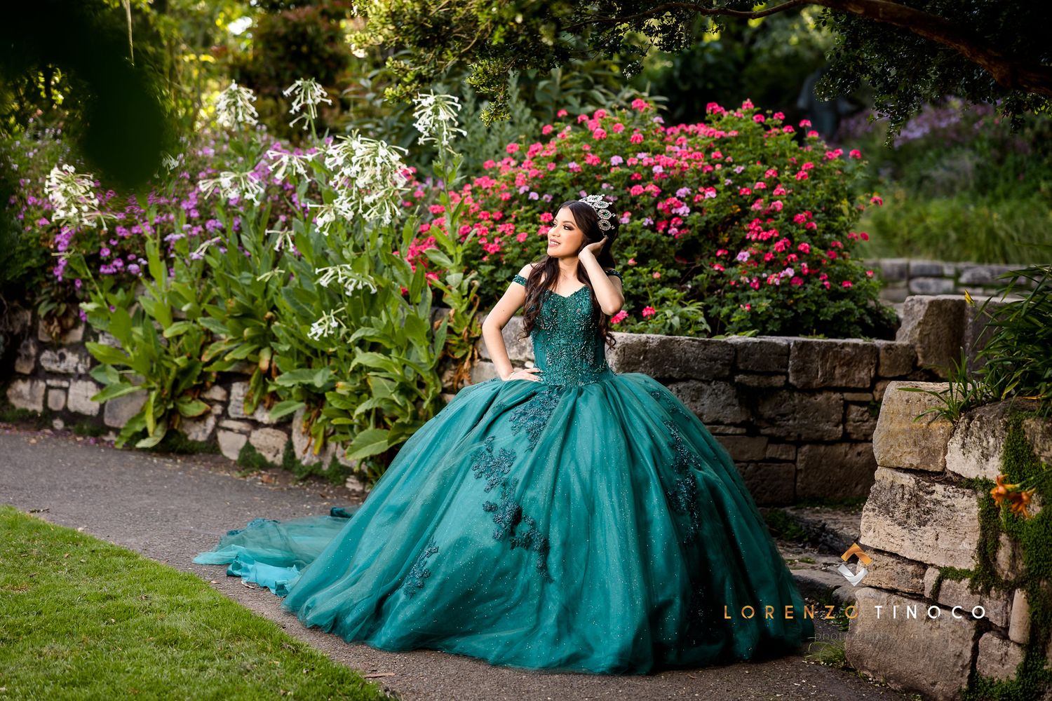 An elegant teal quinceañera dress photographed in a garden setting with blooming flowers and stone walls.