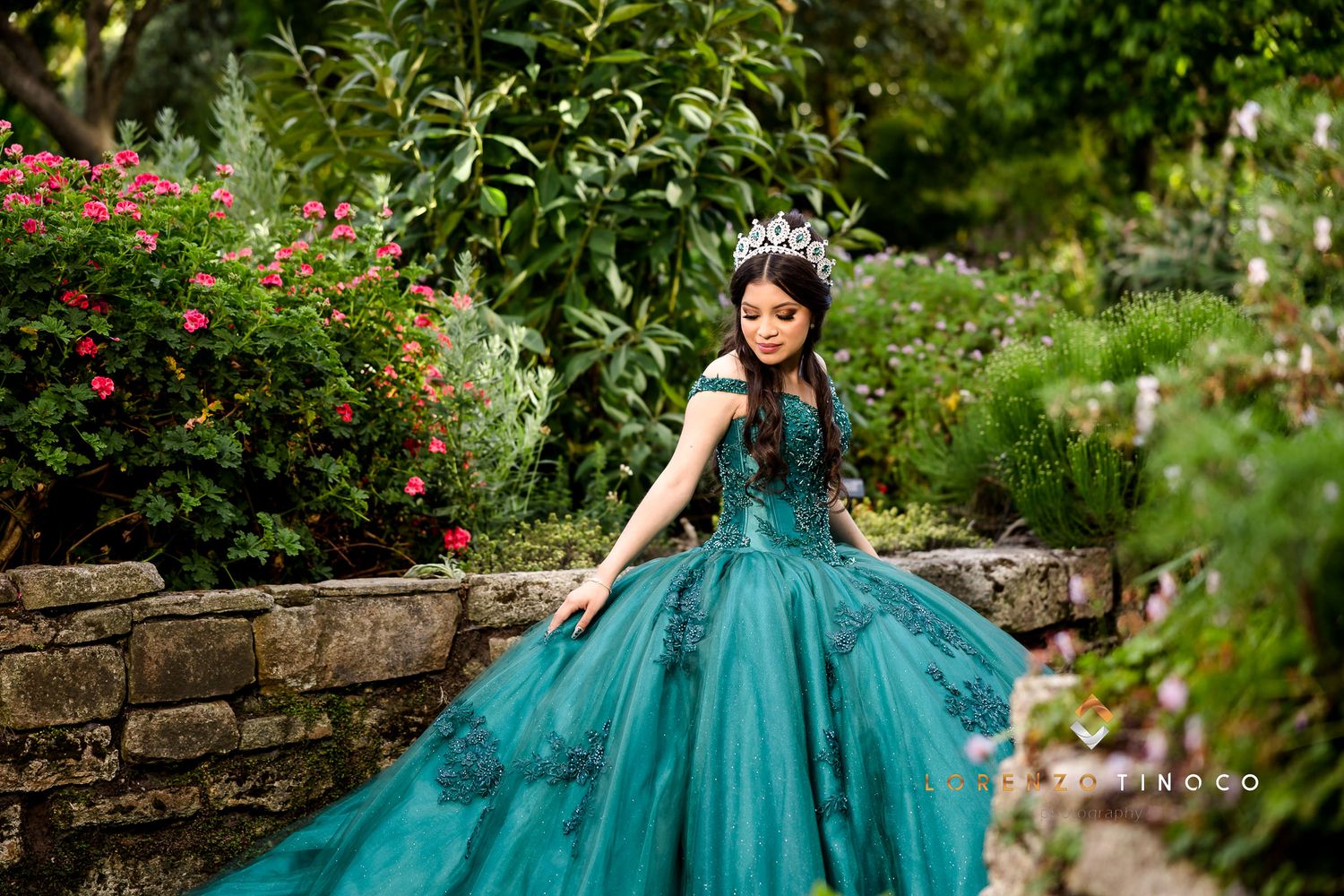 A stunning teal ball gown is displayed in a lush garden setting with red flowering bushes and stone walls.