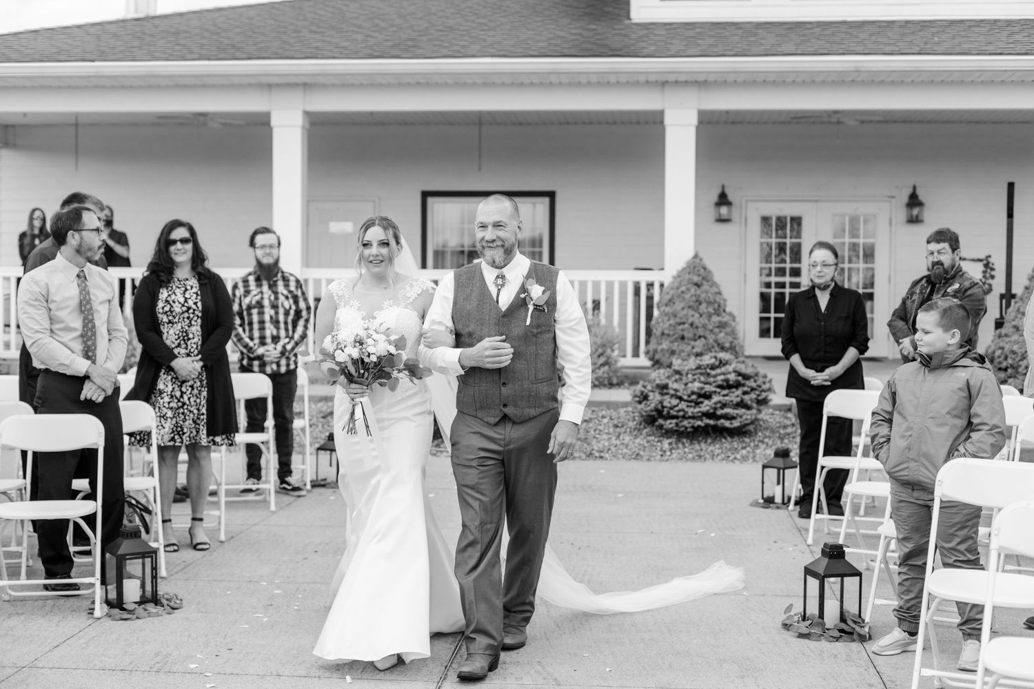 Wedding ceremony guests seated in white chairs on a paved outdoor patio area.