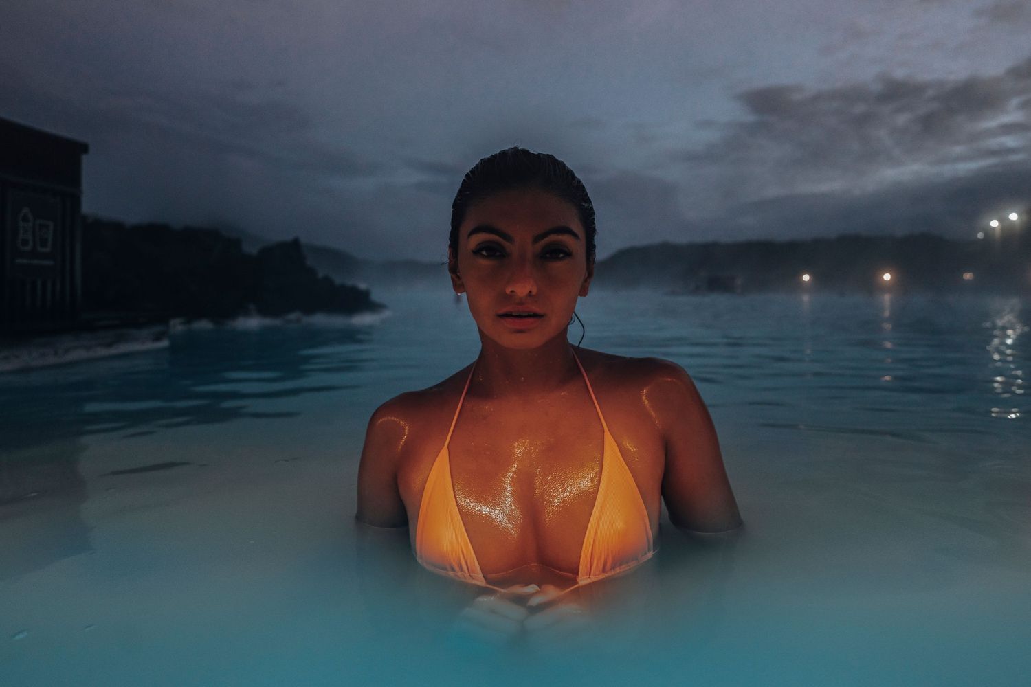 Person bathing in steamy blue geothermal pool at dusk with mountains in background.