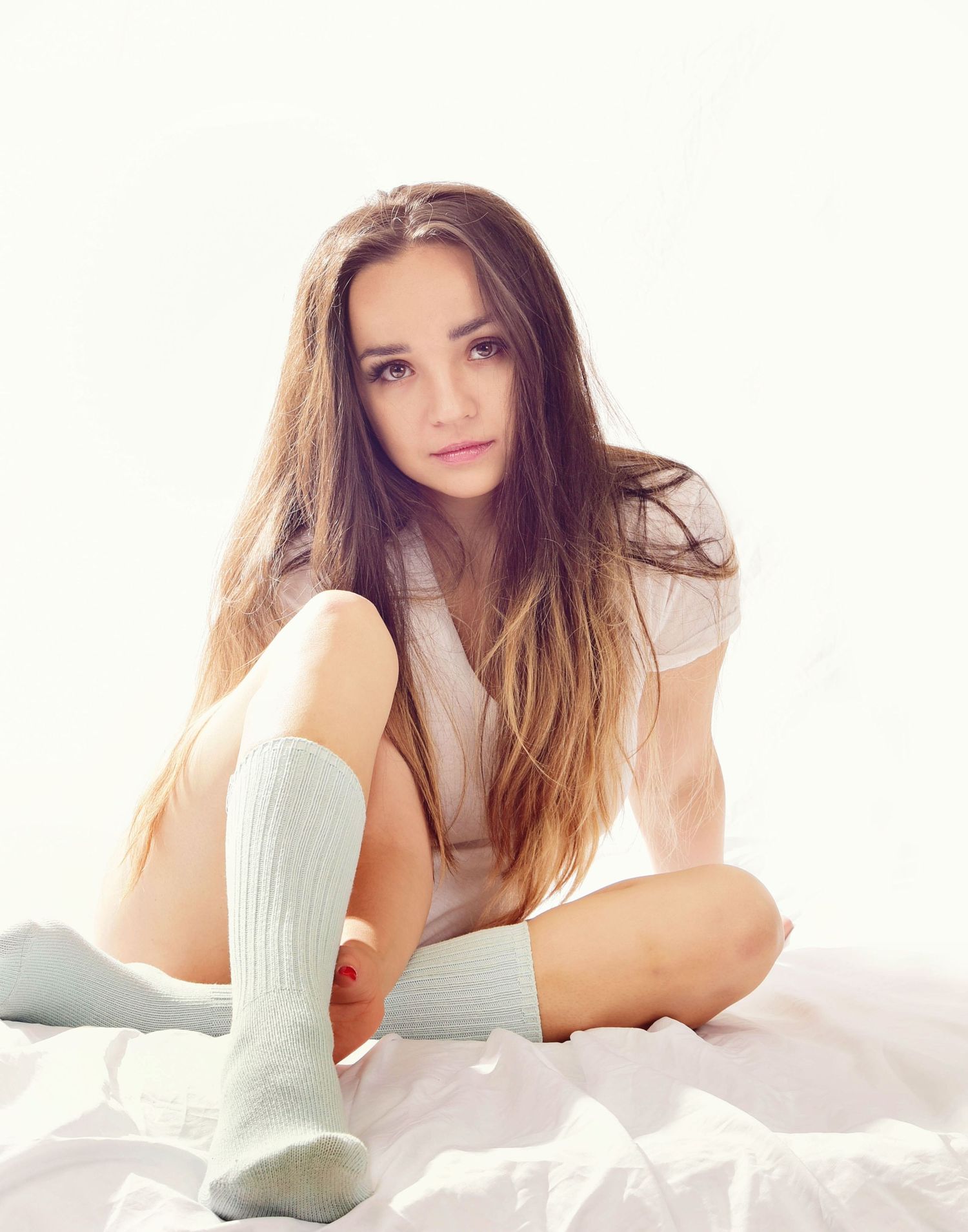 A young woman in long hair and white socks sits on a white surface in a soft, bright setting.