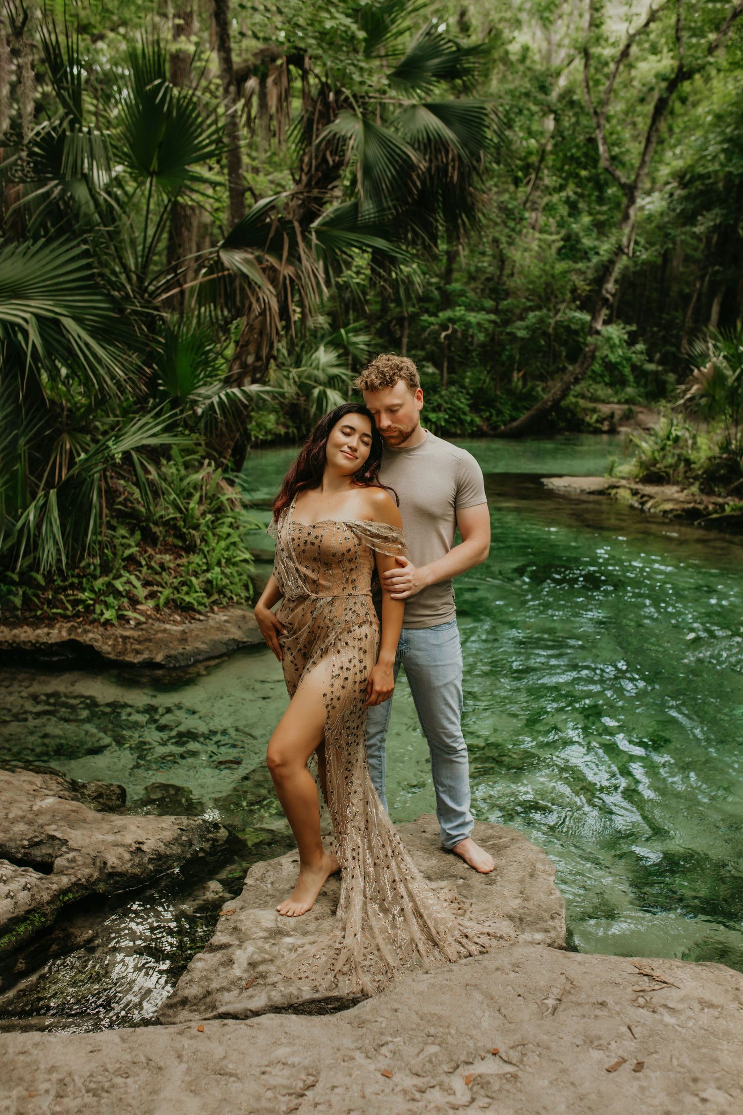 Romantic couple poses in flowing gown beside tropical lagoon with lush palm trees and crystal clear water.