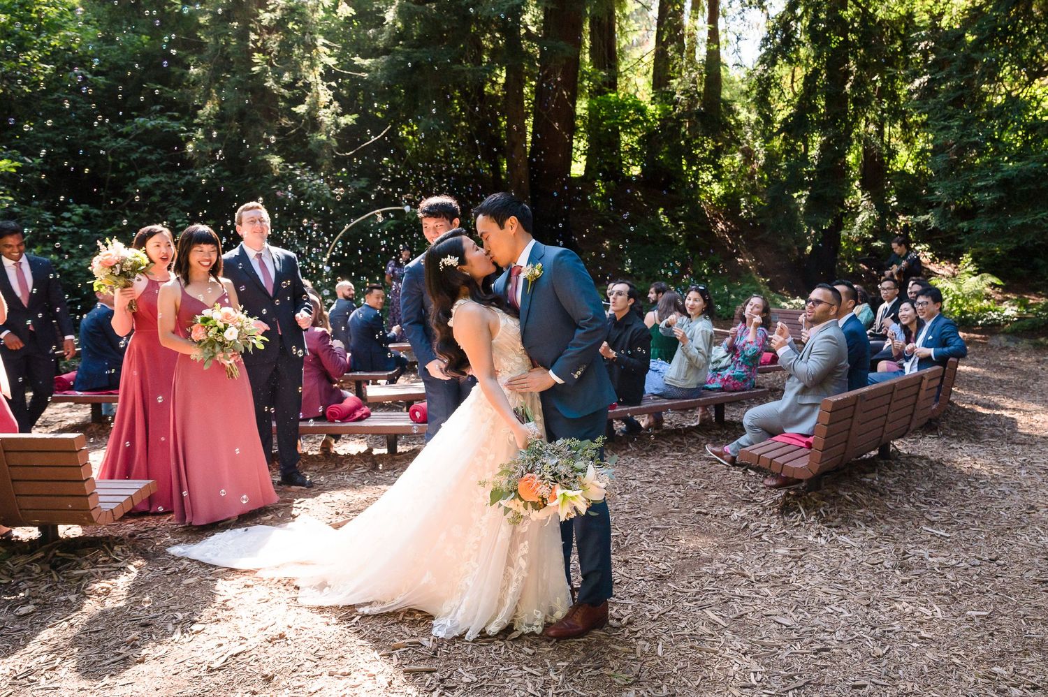 A romantic wedding ceremony in a sunlit forest with guests seated on wooden benches.