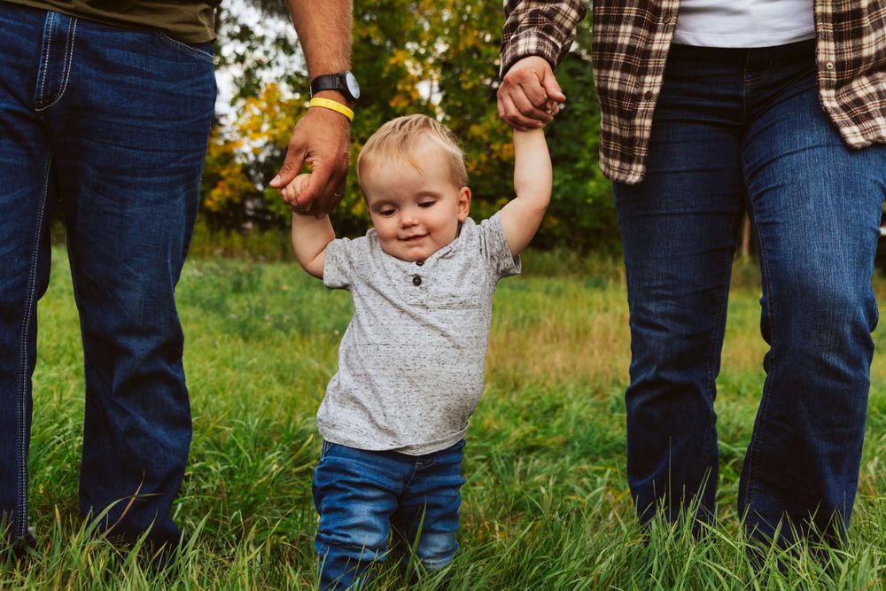 A toddler in a grey shirt holds hands while walking on grass between two people in jeans.