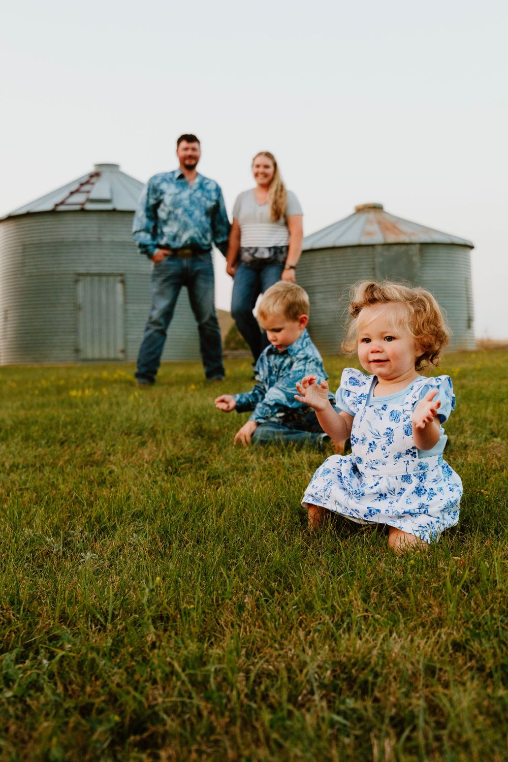Happy family posing for a photo with toddlers on grass near grain silos in countryside setting.