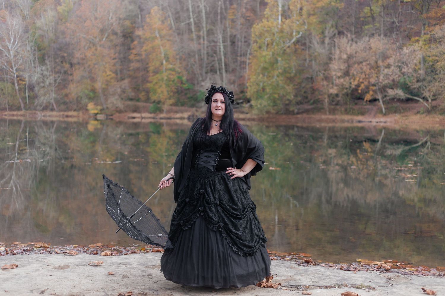 A figure in a black gothic dress poses dramatically by a lake surrounded by autumn foliage.
