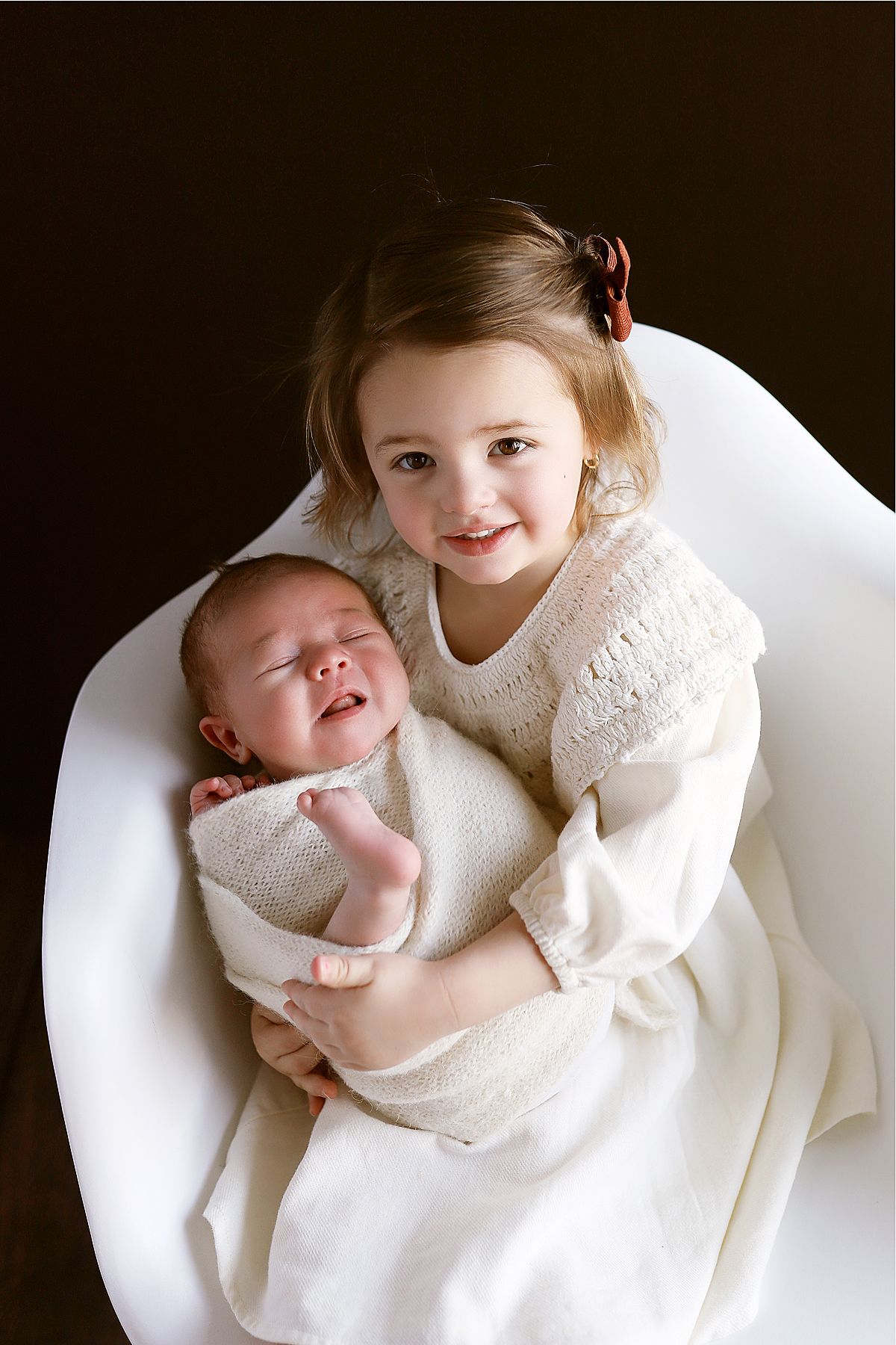 Two children in cream colored outfits share tender moment in white chair.
