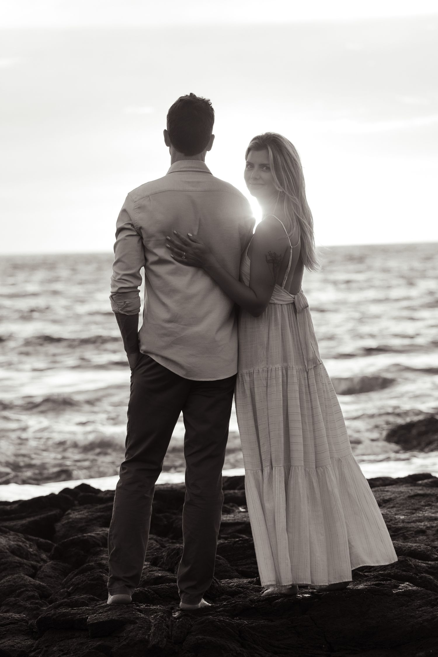 A romantic black and white photo of a couple standing on rocky coastline at sunset.