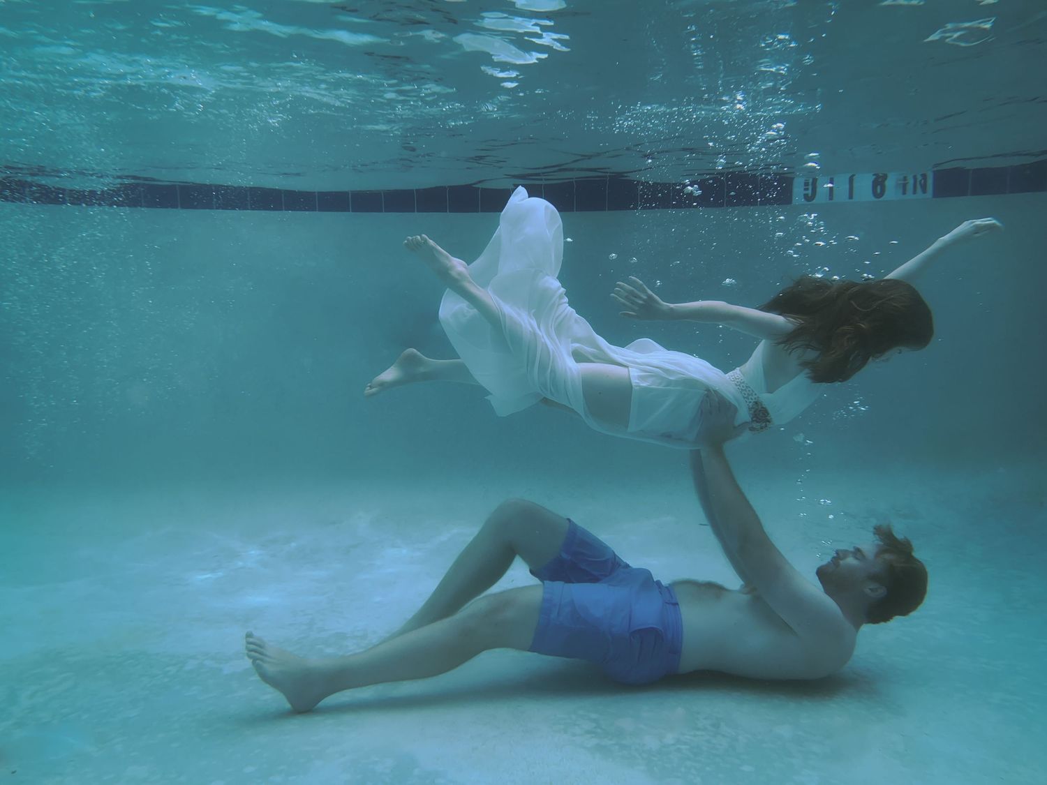 A man lays on the bottom of a pool while holding a woman above him