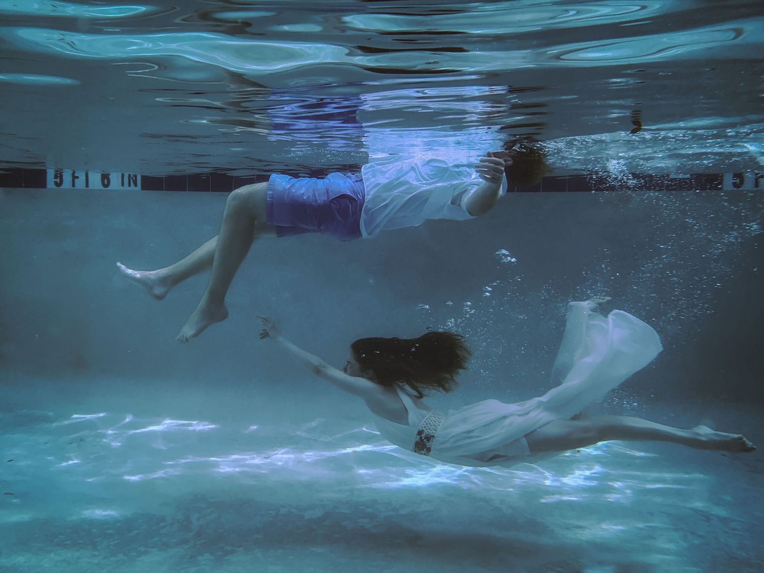 A man swims along the surface of a pool while a woman dives beneath him