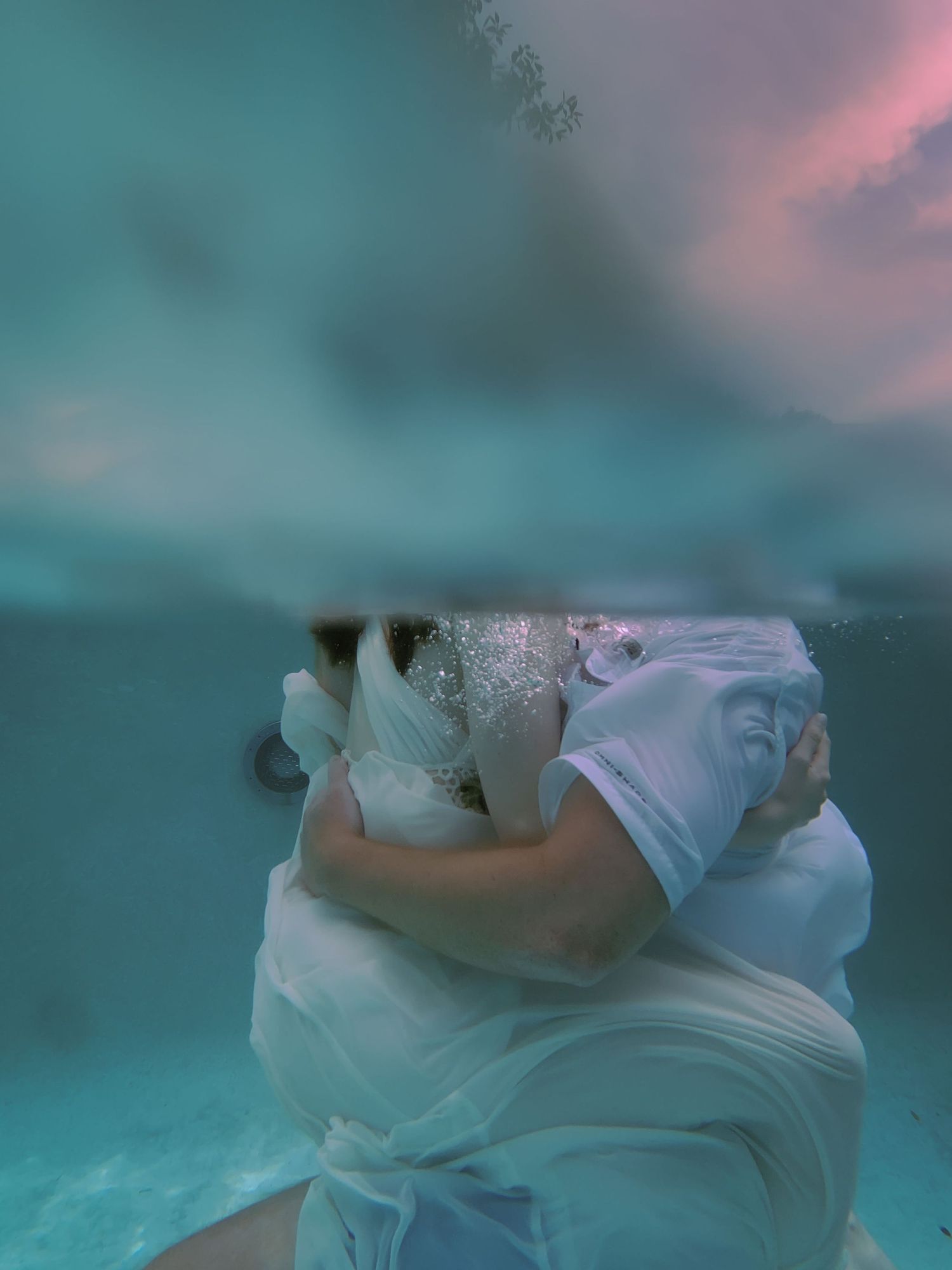A man and woman hold each other under the water while the sunset peeks through