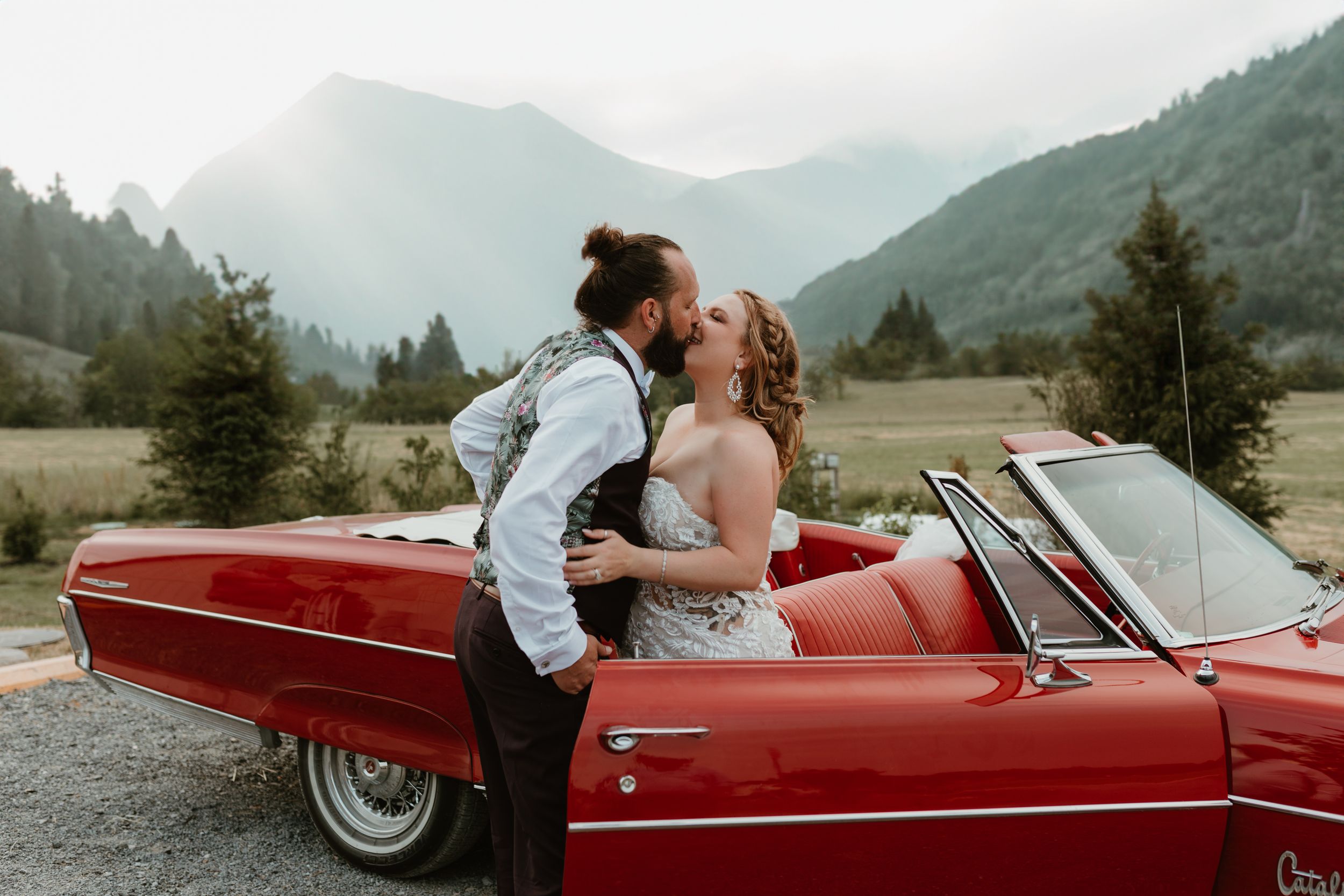 Romantic couple embraces in red vintage convertible car with mountain backdrop.