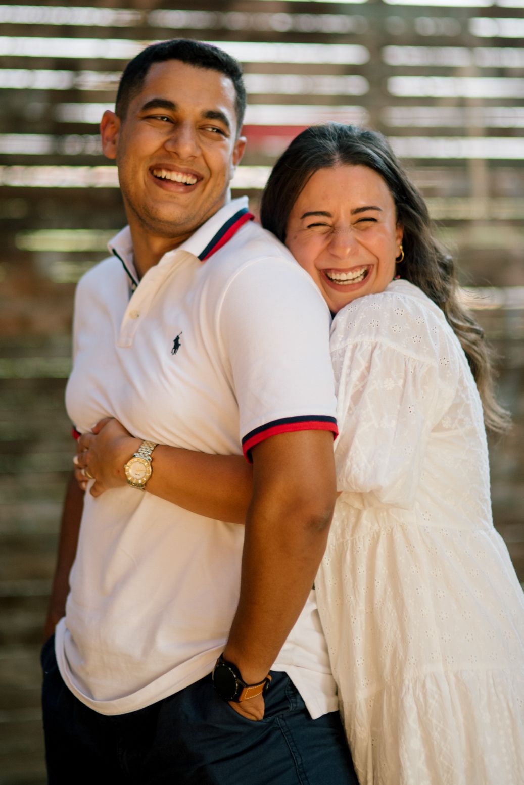 Happy couple laughs together wearing polo and white dress while standing outdoors in warm sunlight.