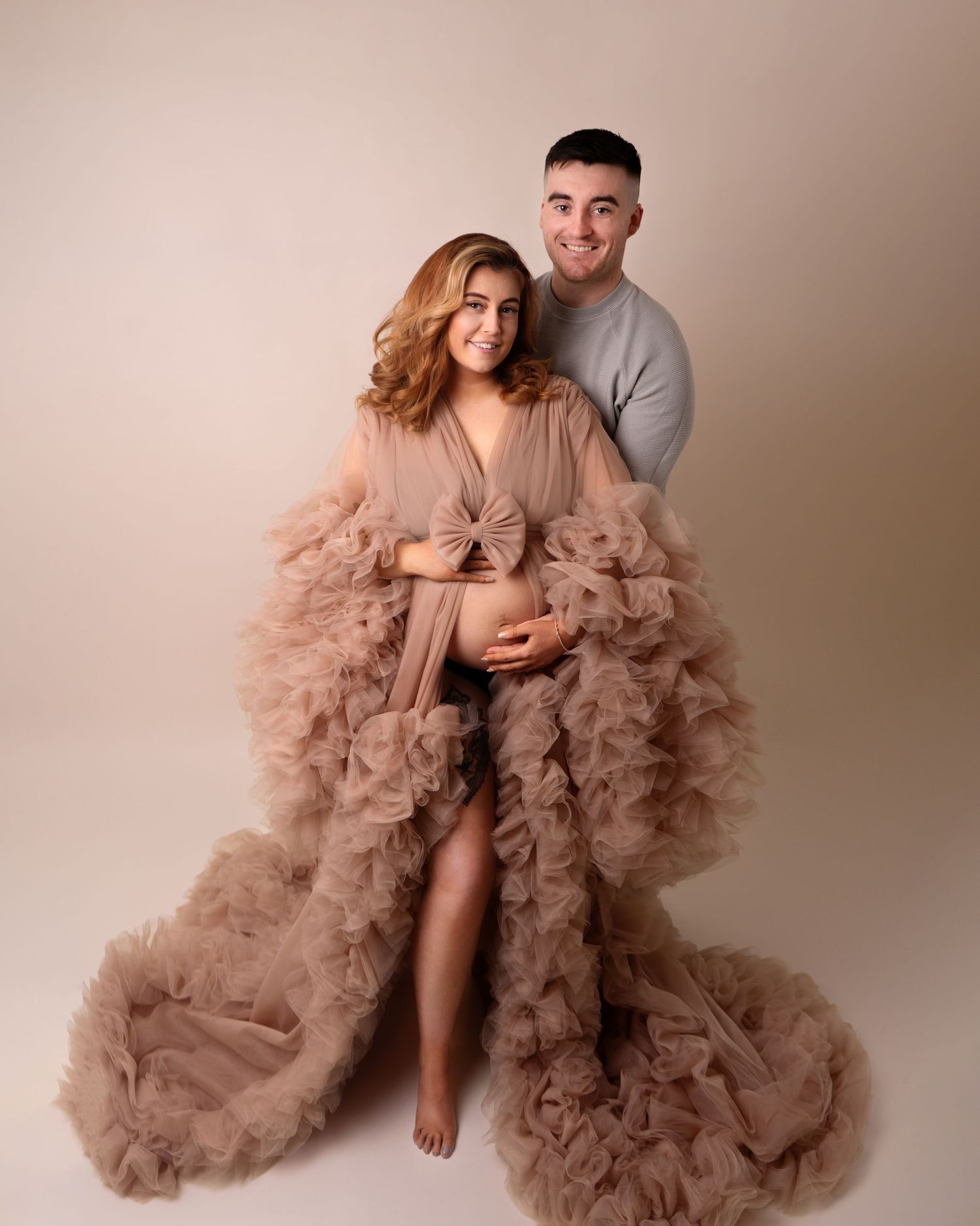 Expectant mother wearing a beige maternity gown holding pregnant bump, with partner at maternity photoshoot in Doncaster