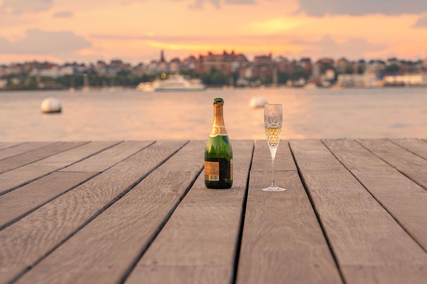 Champagne bottle and glass on wooden deck at sunset with waterfront cityscape view.