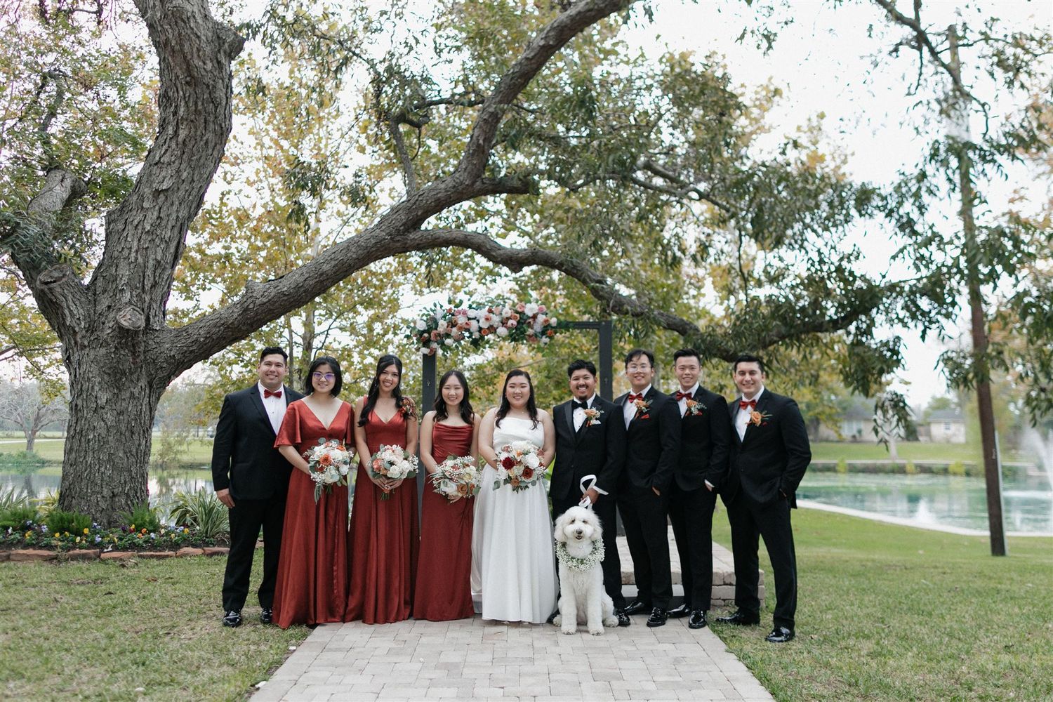 Wedding party in burgundy and black attire poses with couple on stone path under trees.