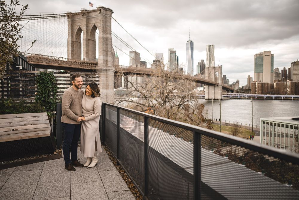 A couple stands on a waterfront terrace with the Brooklyn Bridge and Manhattan skyline in view.