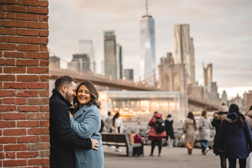 A couple embraces on a brick promenade with city skyline in the background.