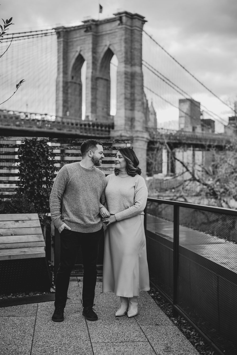 A couple poses on a rooftop terrace with the Brooklyn Bridge visible in the background.