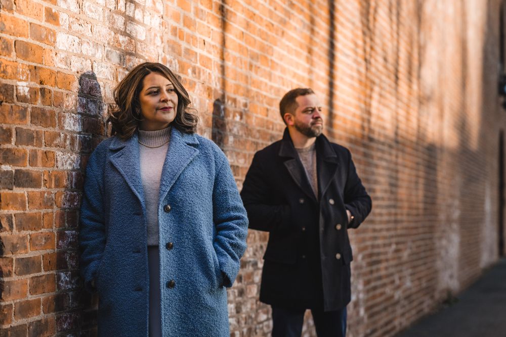 A couple poses against a rustic brick wall in warm sunlight.