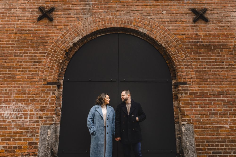 A couple stands in front of an industrial brick wall with black archways.