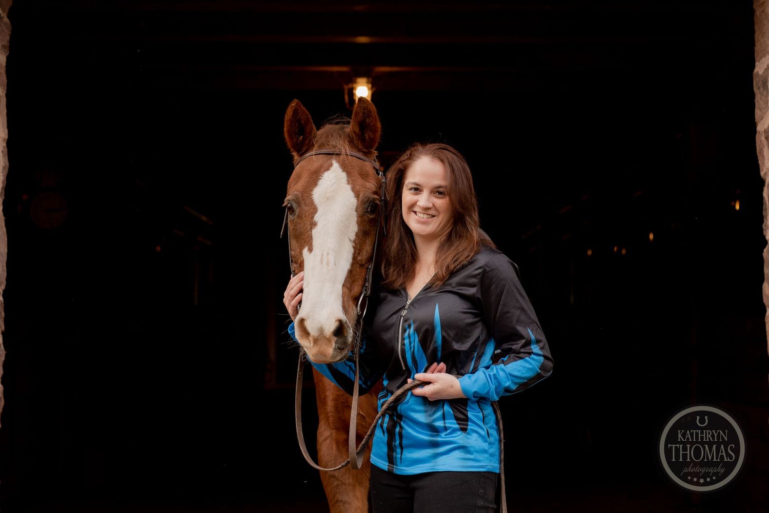 A chestnut horse with white blaze stands in a dark barn doorway beside someone wearing a blue and black jacket.