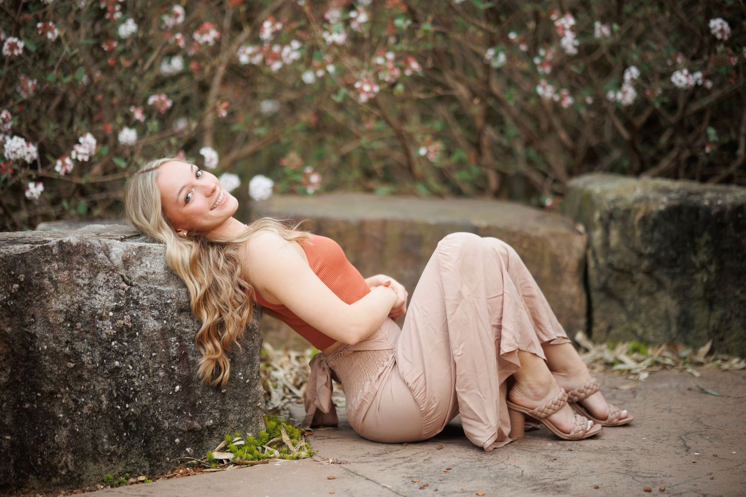 Person in peach dress reclining on stone wall surrounded by flowers.